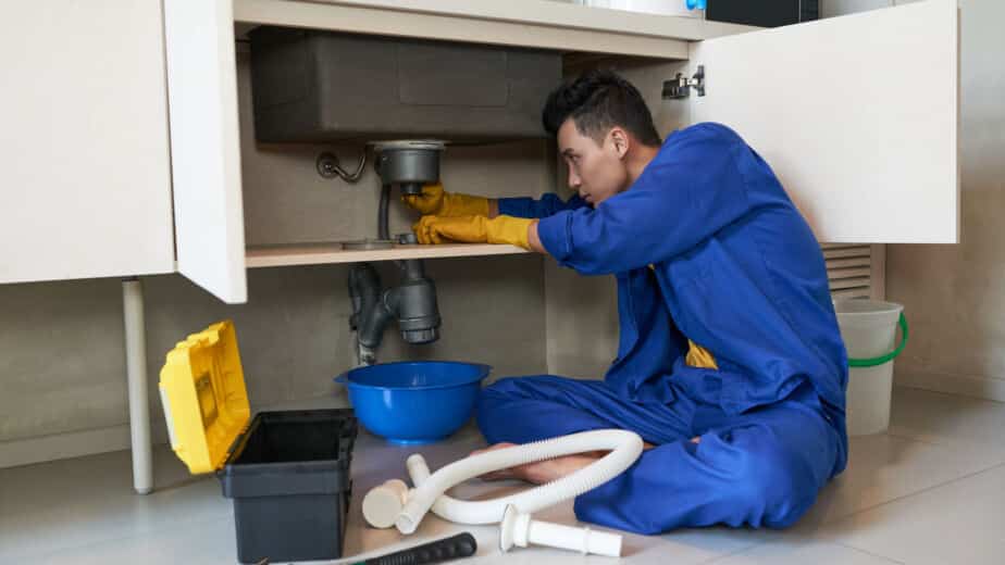 Asian plumber in blue overalls clearing a drain blockage under a sink, surrounded by plumbing tools and equipment, illustrating drain service expertise.