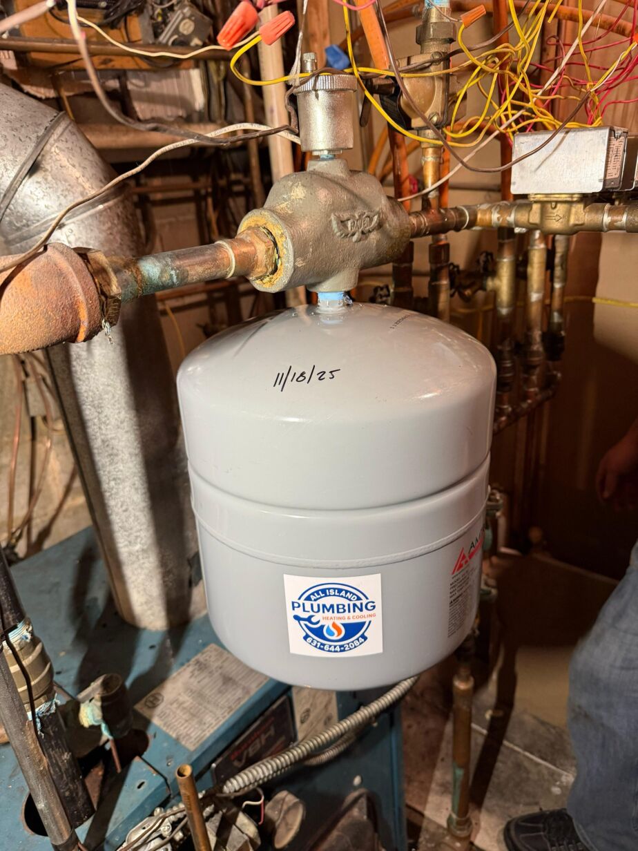 Gray plumbing tank with All Island Plumbing & Heating logo, connected to copper pipes and valves, in a residential heating system setting.