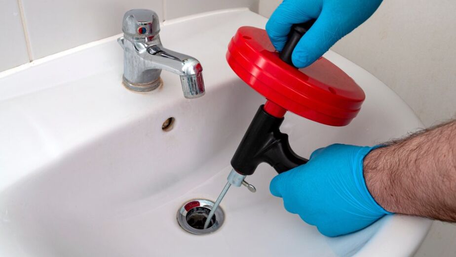 Person using a plumbing snake to clear a clogged sink drain, wearing blue gloves, with a chrome faucet in the background, illustrating drain cleaning services in Nassau County.